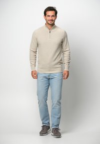 Beige zip-up tröja med en strukturerad finish, bärs över ljusblå jeans och grå sportskor. Tröjan har en ståkrage.