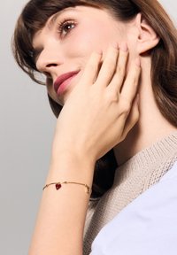 Bransoletka w kolorze złotym z zawieszką w kształcie czerwonego serca, połączona delikatnym łańcuszkiem. Ustawienie ma subtelne, wypolerowane akcenty.