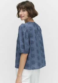 Kortärmad blå blus med broderad blommönster, rund halsringning och avslappnad passform. Den har detaljer med rynkor vid axlarna.