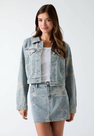 Jeune femme portant une veste en jean acidulé bleu clair et une jupe assortie avec un haut blanc, debout devant un fond clair uni.