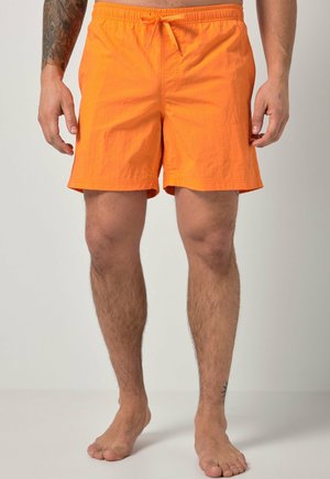 Mężczyzna w jasnopomarańczowych szortach ze sznurkiem, stojący boso na jasnym, jednolitym tle, z tatuażem na lewym przedramieniu.