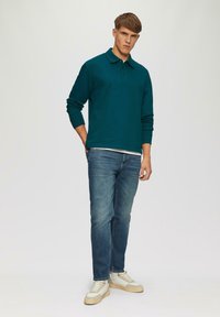 Polo bleu-vert à manches longues avec un col, porté sur un t-shirt blanc, associé à un jean bleu clair et des baskets beiges. Design simple, coupe décontractée.