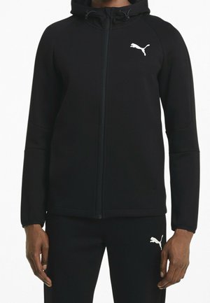 Muž ve černé teplákové soupravě Puma s kapucí na zip a odpovídajícími kalhotami, logo Puma viditelné na hrudi a stehně.
