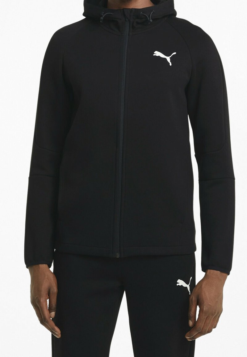 Homme portant un survêtement Puma noir avec sweat à capuche zippé et pantalon assorti, logo Puma visible sur la poitrine et la cuisse.