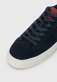 Navy suedesk sneaker med grå snøringer og hvit gummisåle. Har en glatt tekstur og minimal merkevare på siden for et rent design.