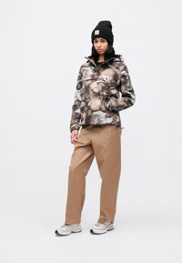 Bedrukte, lichtgewicht jas in tinten bruin en grijs met een buidelzak. Gecombineerd met beige broek en grijze schoenen. Zwarte muts op het hoofd.