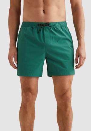 Muž nosiaci zelené plavky s čiernou šnúrkou, stojaci pred jednofarebným svetlošedým pozadím, viditeľný trup a nohy.