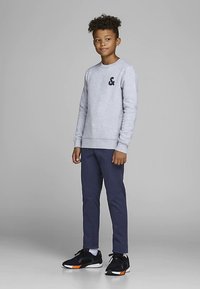 Γκρι φούτερ με μαύρο λογότυπο, σκούρο μπλε παντελόνι και μαύρα αθλητικά παπούτσια με πορτοκαλί λεπτομέρειες. Διαθέτει στρογγυλή λαιμόκοψη και ριμπ μανσέτες.