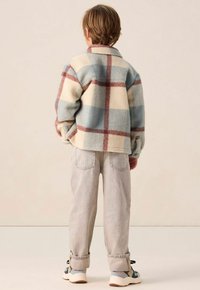 Un enfant vu de dos, portant une veste à carreaux bleu, crème et marron, un pantalon gris clair retroussé, et des baskets multicolores.