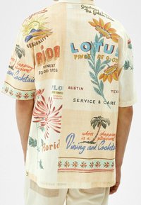 Camisa de manga corta, con botones, en algodón beige. Presenta texto colorido y patrones florales; corte relajado con cuello tipo campamento y una textura suave.