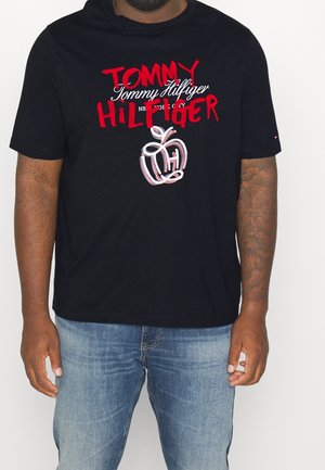 Homme portant un t-shirt noir Tommy Hilfiger avec un logo rouge et blanc et un motif graphique, assorti à un jean bleu, debout devant un fond blanc.