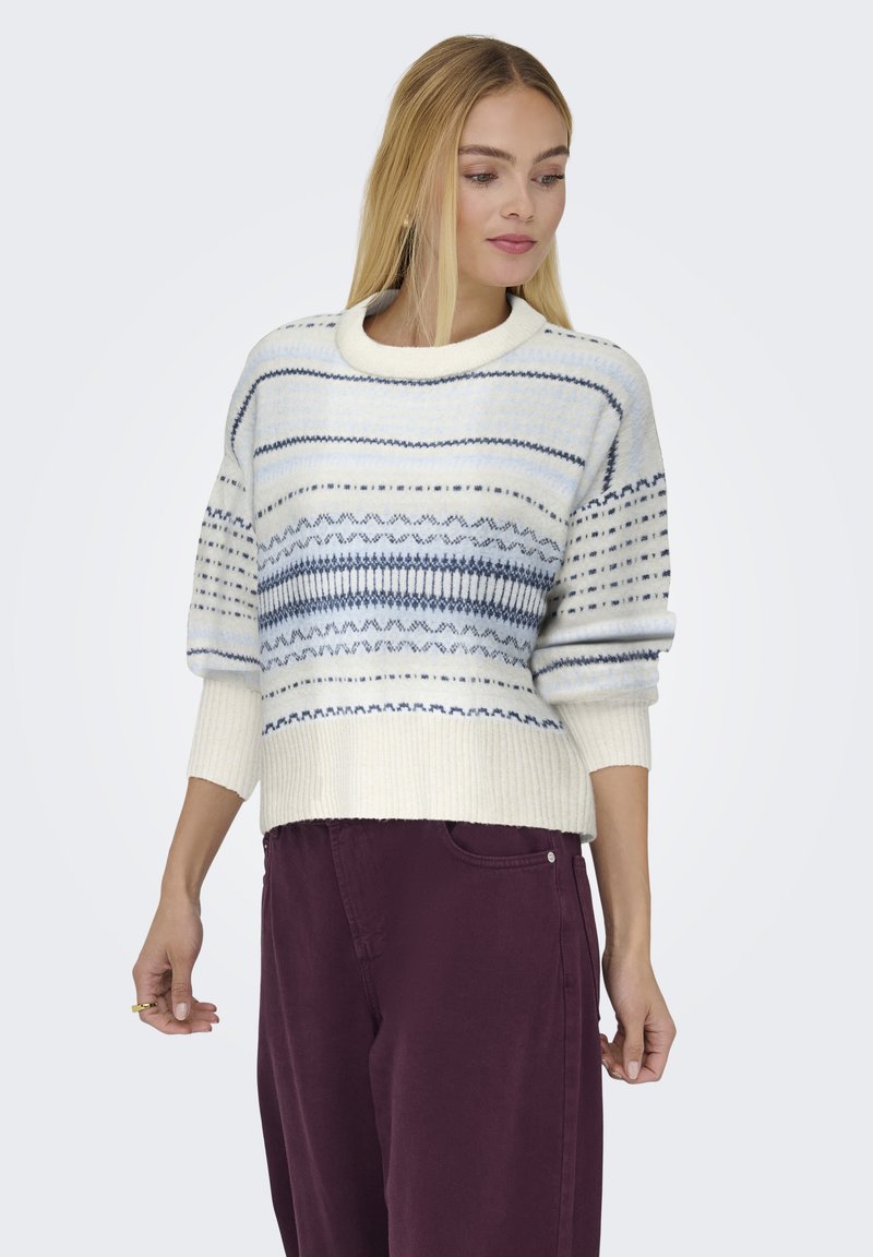 Strickpullover in Creme mit blauen Mustern, Rundhalsausschnitt, verkürztem Design und gerippten Bündchen, kombiniert mit burgunderfarbenen Hosen.