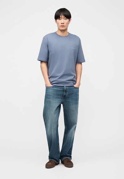 Άνδρας που στέκεται με τα χέρια στις τσέπες, φορώντας μπλε υπερμεγέθη μπλούζα, φαρδιά μπλε τζιν και καφέ αθλητικά παπούτσια, μπροστά από απλό φόντο.