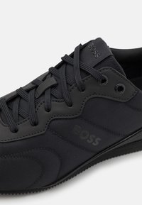 Sort sneaker med et tekstureret stofoverdel, glatte syntetiske accenter, tonede snørebånd og et præget logo på siden.