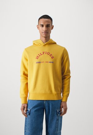 Hoodie giallo in misto cotone, con testo rosso "HILFIGER" e "EST. 1985" sotto, e strisce a contrasto. Tasca a marsupio frontale.