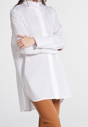 Femme portant une chemise blanche ample à manches longues boutonnée, les bras croisés, et un pantalon marron slim, debout devant un fond uni.