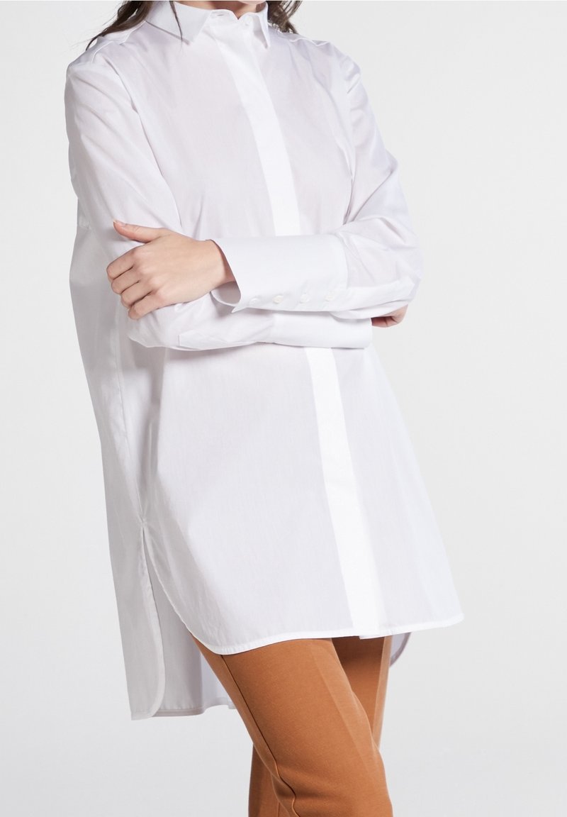 Femme portant une chemise blanche ample à manches longues boutonnée, les bras croisés, et un pantalon marron slim, debout devant un fond uni.