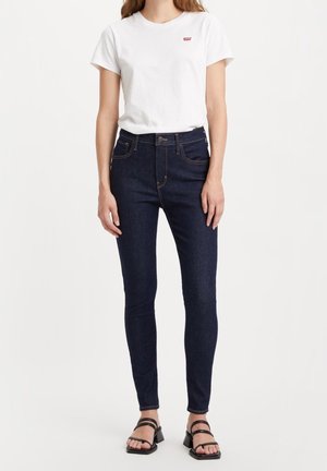 Dark blue skinny jeans paired with a plain white t-shirt featuring a small red logo. The jeans have front pockets and a button closure.