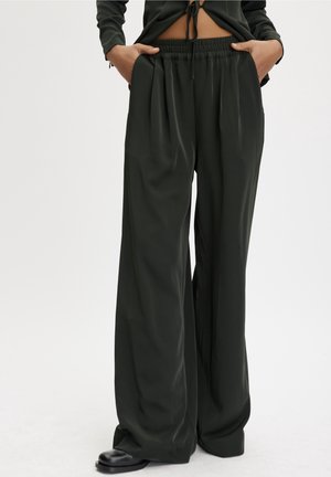 Persoon die zwart, hooggetailleerd, wijd uitlopende broek draagt met elastische tailleband en handen in de zakken, gecombineerd met zwarte schoenen en een zwart, kort topje.