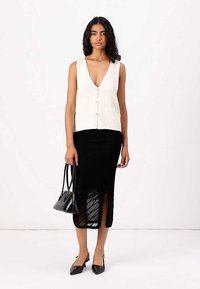 Biała bezrękawnikowa bluzka z dekoltem w kształcie litery V i guzikami z przodu, zestawiona z czarną, półdługą przezroczystą spódnicą z bocznym rozcięciem. Czarna torebka i szpilki w szpic.