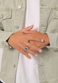 Anello d'argento con una pietra striata verde poggia su delle dita, abbinato a una giacca di jeans verde chiaro sopra una camicia bianca. Messa a fuoco ravvicinata.