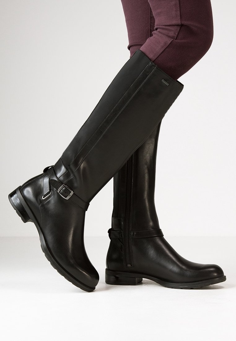 Botas de cuero negro hasta la rodilla con cremallera lateral, detalle de correa y hebilla, punta redondeada y acabado texturizado, combinadas con pantalones de color púrpura oscuro.