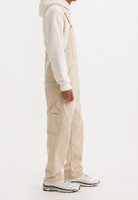 Peto de pana beige con rayas verticales, que cuenta con un bolsillo frontal, tirantes ajustables y un corte holgado, combinado con zapatillas blancas.