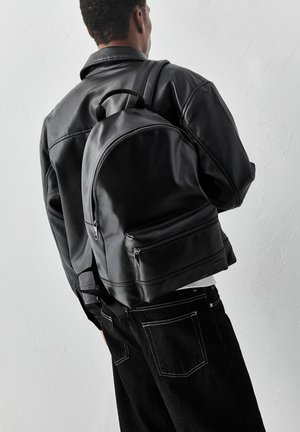 Személy fekete bőrkabátban és fekete hátizsákkal, háttal egy sima világos falnak.