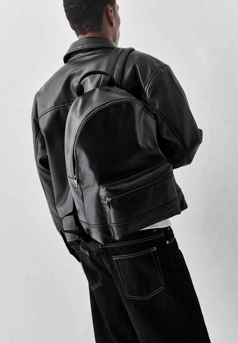 Osoba v čiernej koženej bunde a s čiernym batohom, otočená chrbtom k jednoduchej svetlej stene.