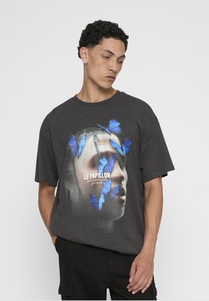 Junger Mann trägt ein dunkelgraues T-Shirt mit einem Gesicht und mehreren blauen Schmetterlingen, mit dem Schriftzug „Le Papillon“ in der Mitte.