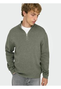 Zielony sweter na zamek z wysokim kołnierzem, z prążkowaną fakturą i długimi rękawami, nałożony na białą koszulę, w połączeniu z szarymi dżinsami.