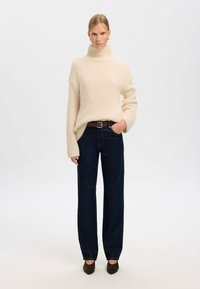 Pulover bej tricotat cu guler înalt și mâneci cu model rebatut, asortat cu blugi largi de culoare albastru închis și pantofi cu toc de culoare verde închis.