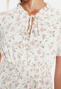 Blommig klänning med ett vitt tyg med rosa och gröna blomstertryck, nyckelhålsringning med knytningar och elastisk midja.