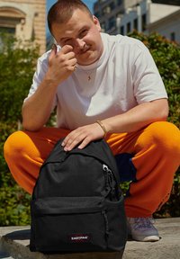 Mochila negra con una tela texturizada, múltiples bolsillos con cremallera y un parche con logo. El usuario lleva una camiseta blanca de gran tamaño y pantalones naranjas.