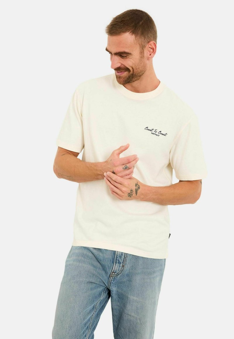 T-shirt en coton couleur crème à manches courtes et col rond, avec une broderie noire "Coast To Coast" sur le côté gauche de la poitrine.