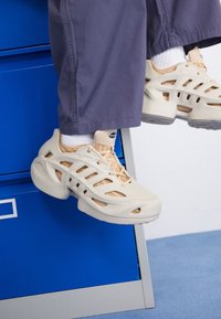 Kengät beigen ja vaaleanruskean suunnittelun kanssa, joissa on leikattuja paneeleita, teksturoitua kangasta ja nauhakiinnitys, sijoitettuna siniselle pinnalle.