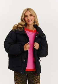 Žena s blond vlasy, oblečená v černé prošívané bundě přes růžový svetr s červenými detaily a kalhoty s leopardím vzorem, usmívající se na jednobarevném pozadí.
