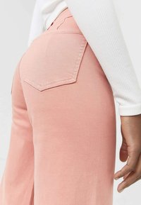 Pantaloni a gamba larga rosa chiaro con una texture liscia, dotati di una tasca posteriore con contorno cucito. Una maglietta bianca a maniche lunghe ben aderente è parzialmente visibile.