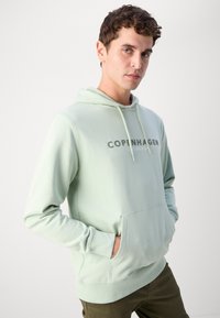 Felpa verde chiaro realizzata in tessuto morbido, con tasca frontale e cappuccio con cordino. La scritta "COPENAGHEN" è ben visibile sul petto.
