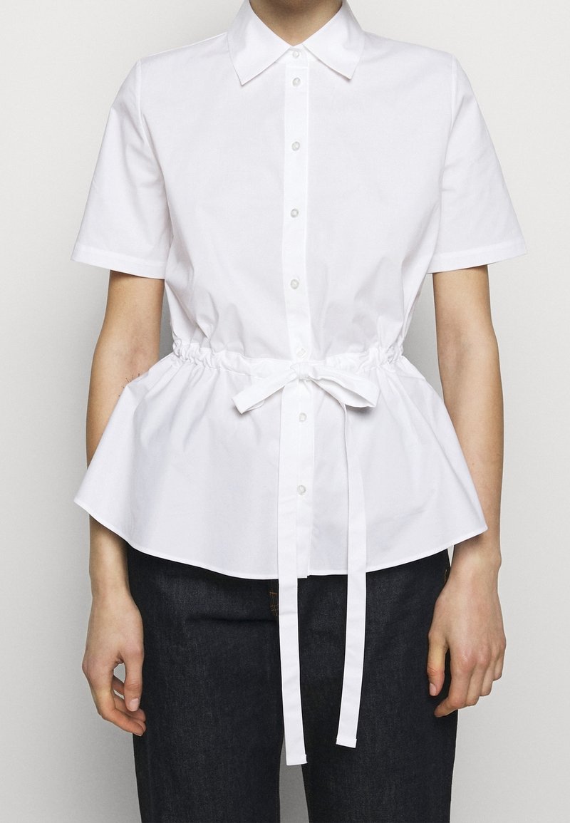Chemise en coton blanc avec un corsage ajusté, des manches courtes et un ourlet à volant, dotée d'un lien à cordon de serrage à la taille et d'une fermeture à boutons.