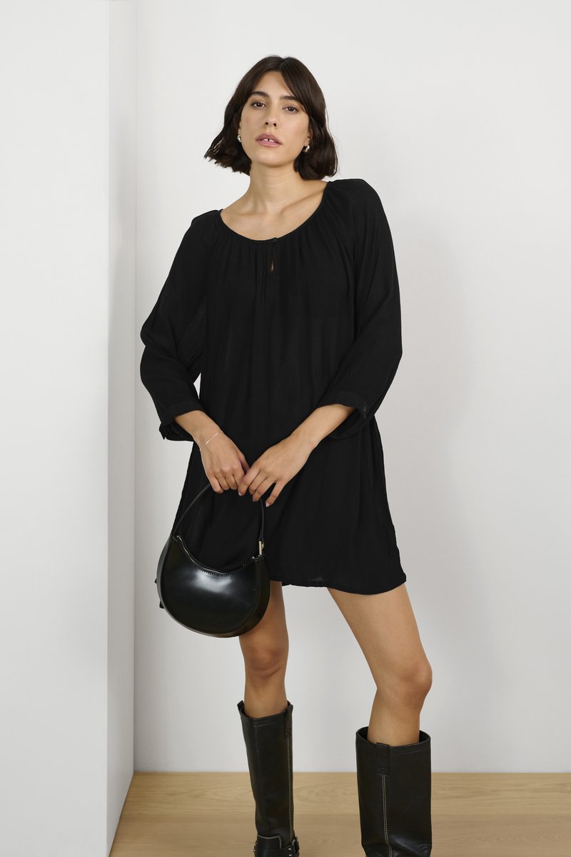 Schwarzes langärmliges Kleid mit einem Schlüssellochnacken und strukturiertem Stoff, kombiniert mit einer kleinen schwarzen runden Handtasche und kniehohen schwarzen Stiefeln.