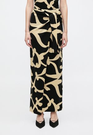 Mulher usando uma saia longa preta com grandes padrões abstratos bege, com detalhe atado na cintura, combinada com sapatos pretos de bico pontudo.