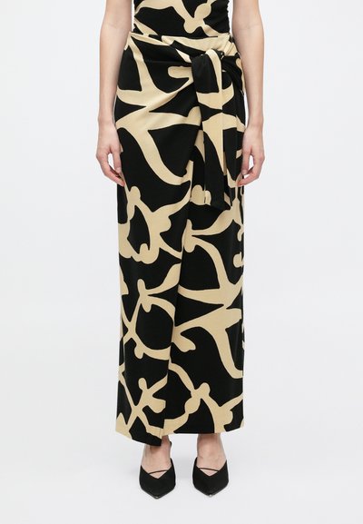 Femme portant une longue jupe noire avec de grands motifs abstraits beiges, détail noué à la taille, accompagnée de chaussures noires à bout pointu.