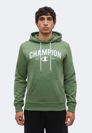 Junger Mann trägt grünen Champion-Hoodie mit weißem Logo und schwarzen Hosen, steht vor einfarbig hellem Hintergrund.