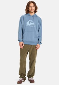 Světle modrá mikina s kapucí a velkým bílým logem Quiksilver. Má klokání kapsu a kapuci se stahovací šňůrkou. Nošena s olivově zelenými kalhotami.