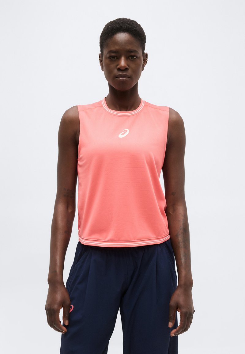 Femme portant un haut de sport corail sans manches et un pantalon bleu marine, debout face à l’avant avec une expression neutre.