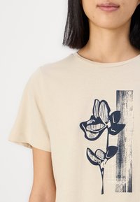 Beige bavlnené tričko s krátkymi rukávmi, s námornícky modrým kvetinovým grafickým dizajnom a vertikálnym vzorom na prednej strane, okrúhly výstrih.