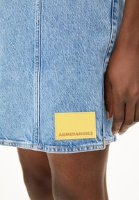 Lichtblauwe denim rok met een geel stoffen label waarop "ARMEDANGELS" in bruine tekst staat. Close-up op de stiksels en labeldetails.