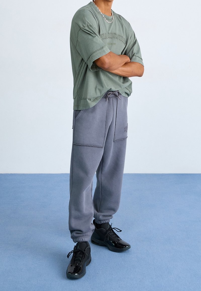 Homme portant un t-shirt vert ample, un pantalon de survêtement gris avec de grandes poches, des baskets noires et une chaîne argentée, debout sur un sol bleu les bras croisés.