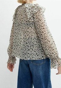 Blouse transparente à imprimé floral noir et blanc, col à volants et manches ballon, associée à un jean en denim bleu.